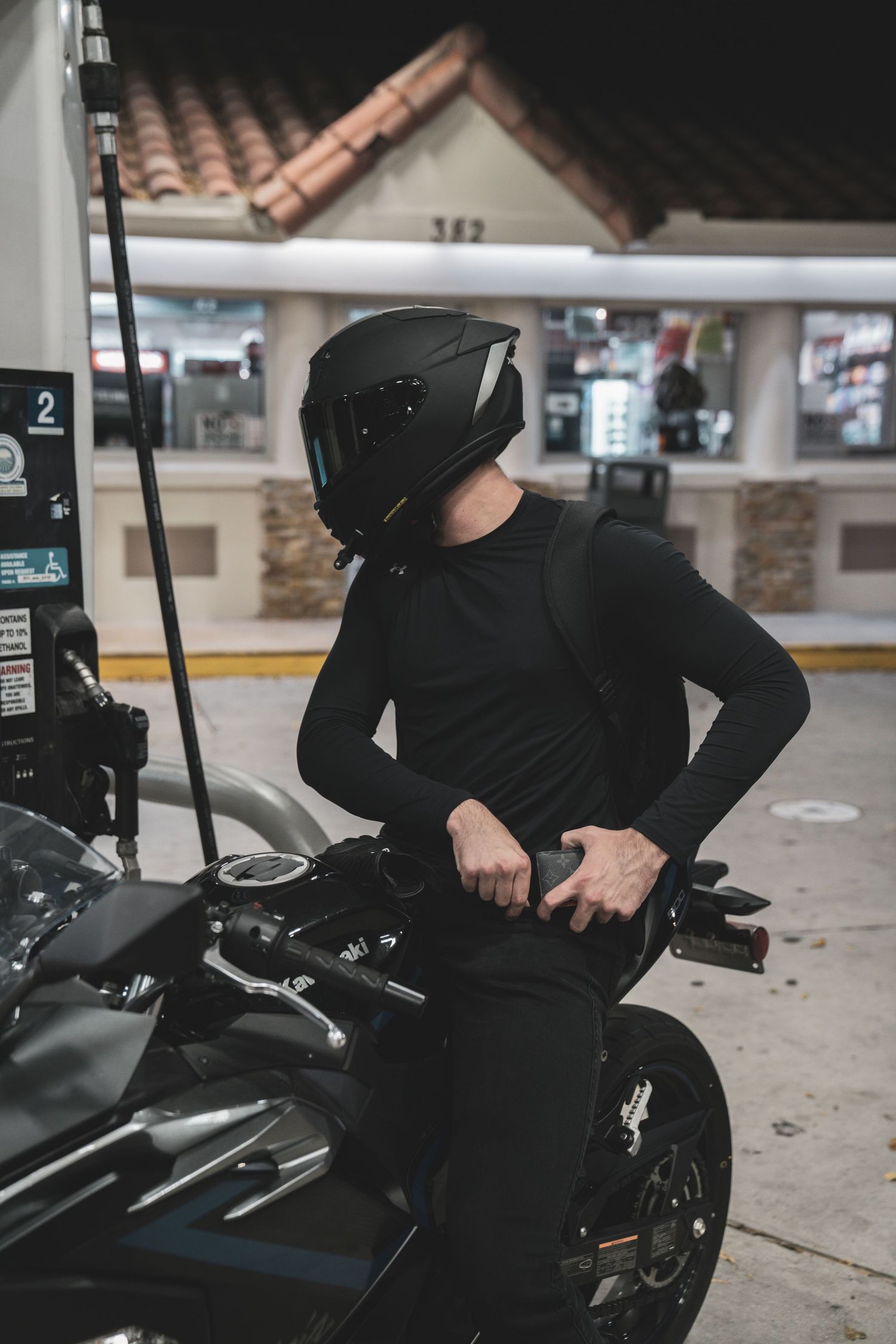 A motorcyclist in black gear sits on a parked motorcycle in an urban setting.