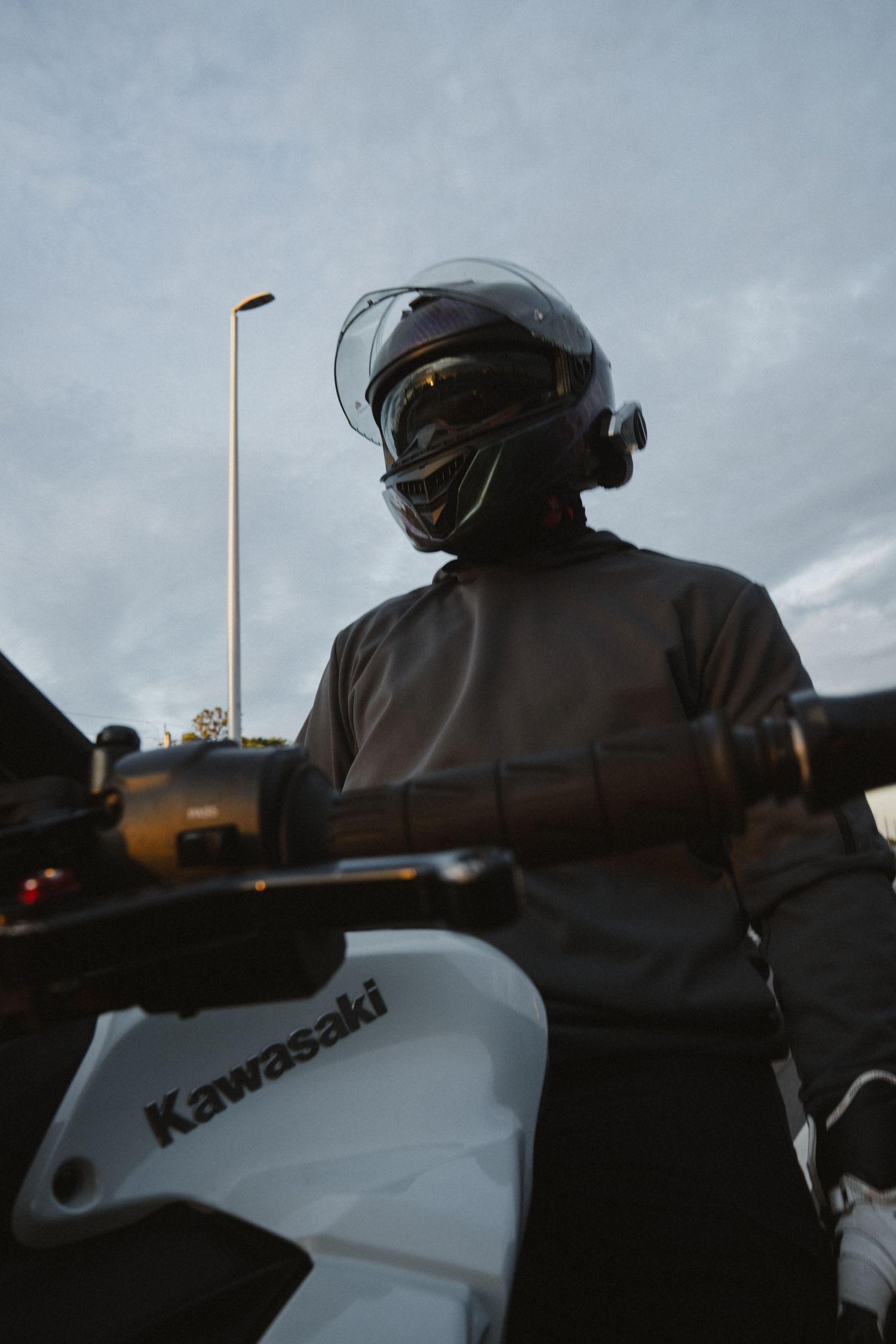 Motorcyclist in full-face helmet riding a white Kawasaki motorcycle against a cloudy sky backdrop.