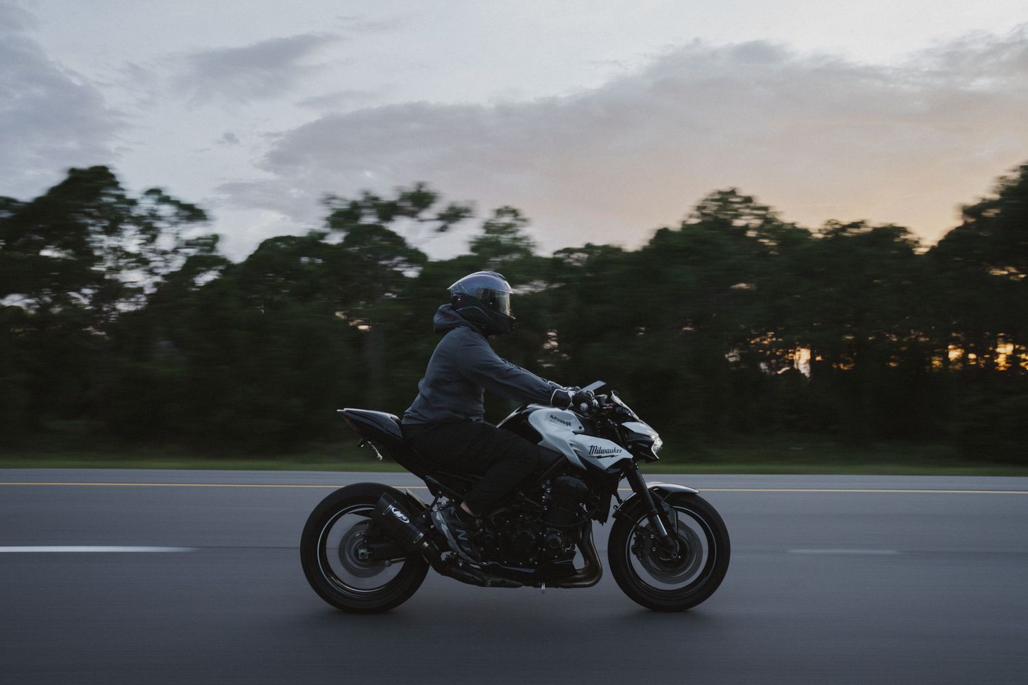 Motorcyclist riding a black motorcycle on a highway at dusk with trees and cloudy sky in the background.