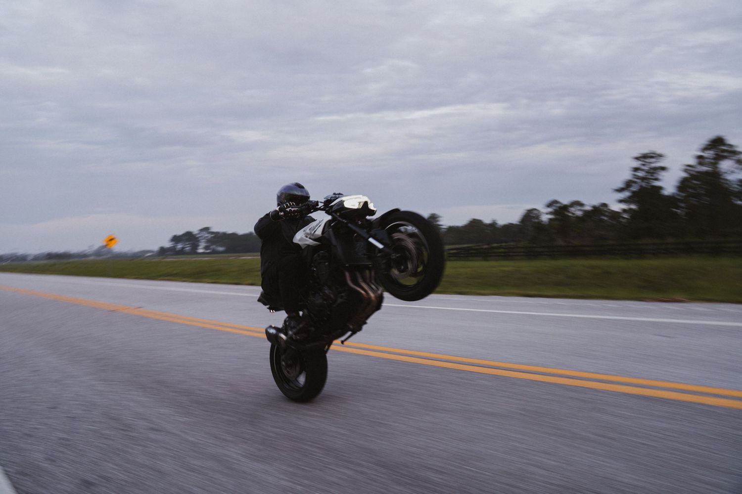Motorcyclist performing a wheelie on a wet road with trees and cloudy sky in the background.