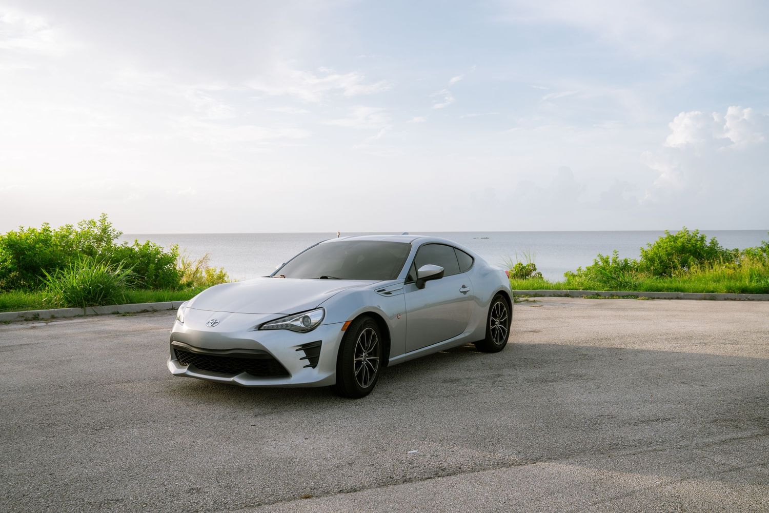 Silver Toyota 86 sports car parked on pavement near ocean with lush greenery and cloudy sky in background.