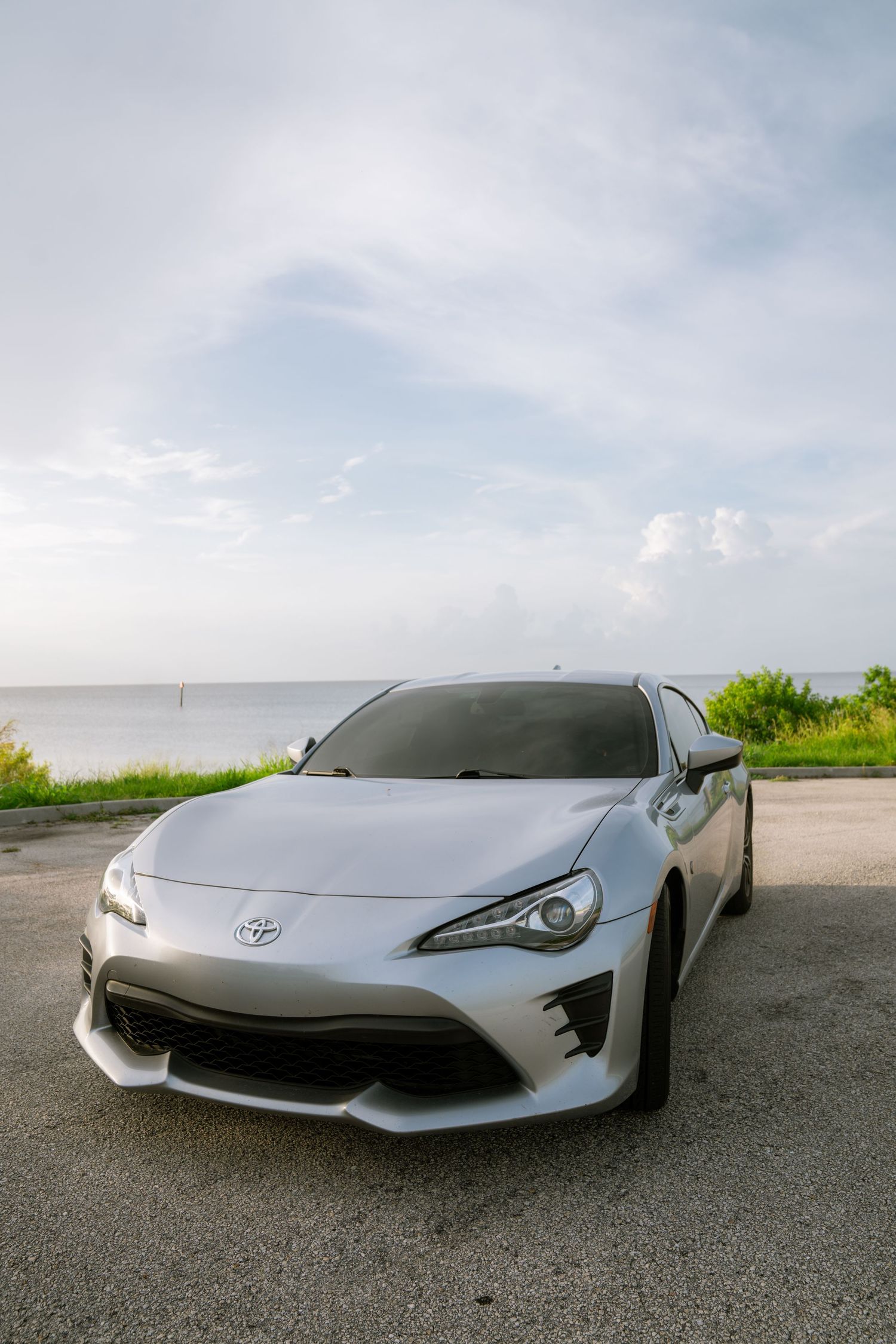 White Toyota 86 sports car parked near oceanfront with cloudy sky backdrop.