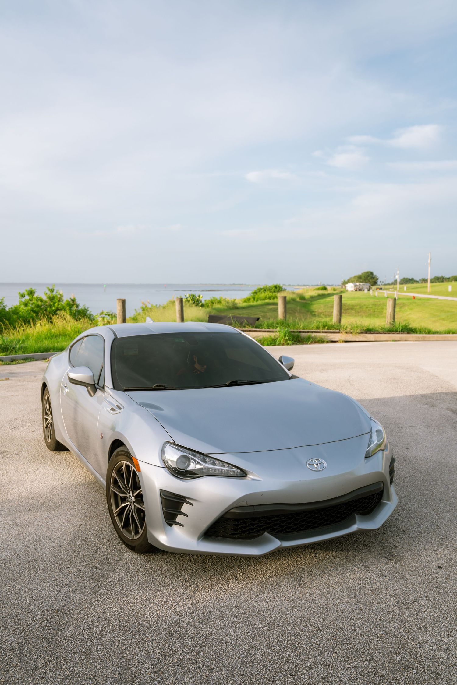 Silver Toyota 86 sports car parked near waterfront on sunny day with green grass and blue sky background.