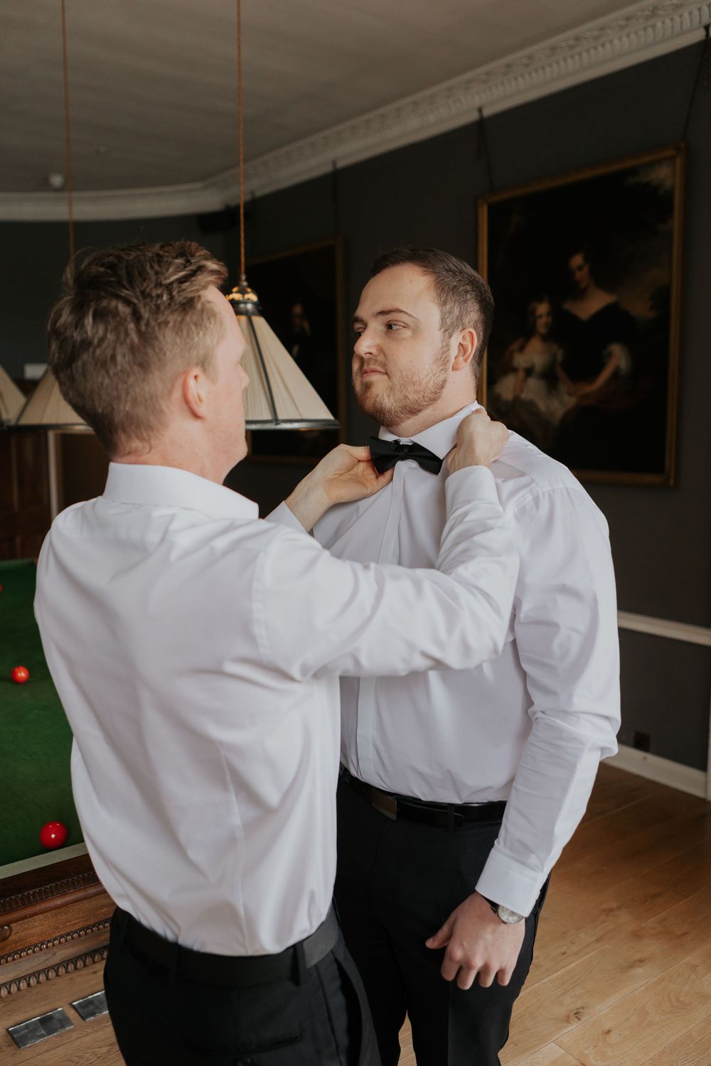 Two men in white shirts, one adjusting the other's black bow tie before a formal event.