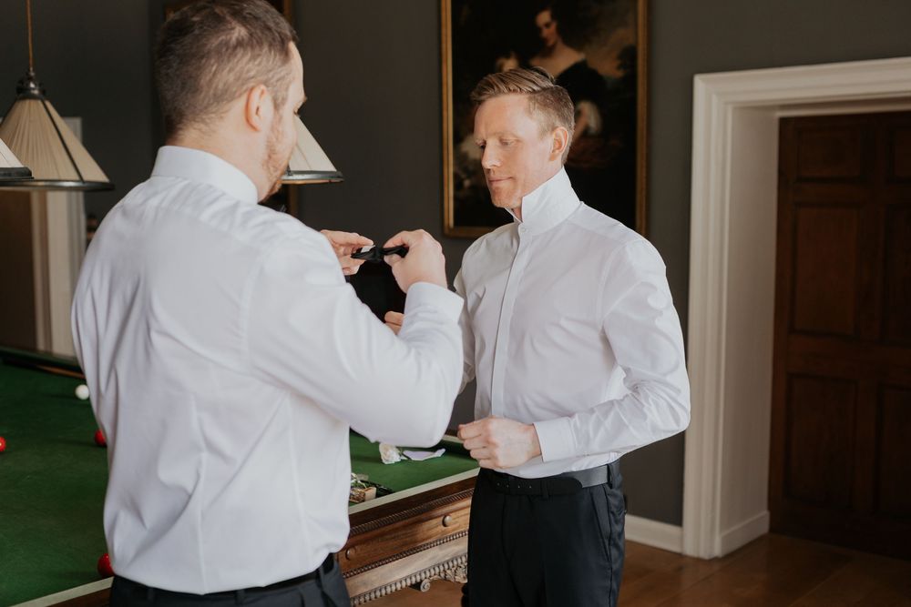 Two men in white shirts helping each other prepare in a room with a green billiard table.