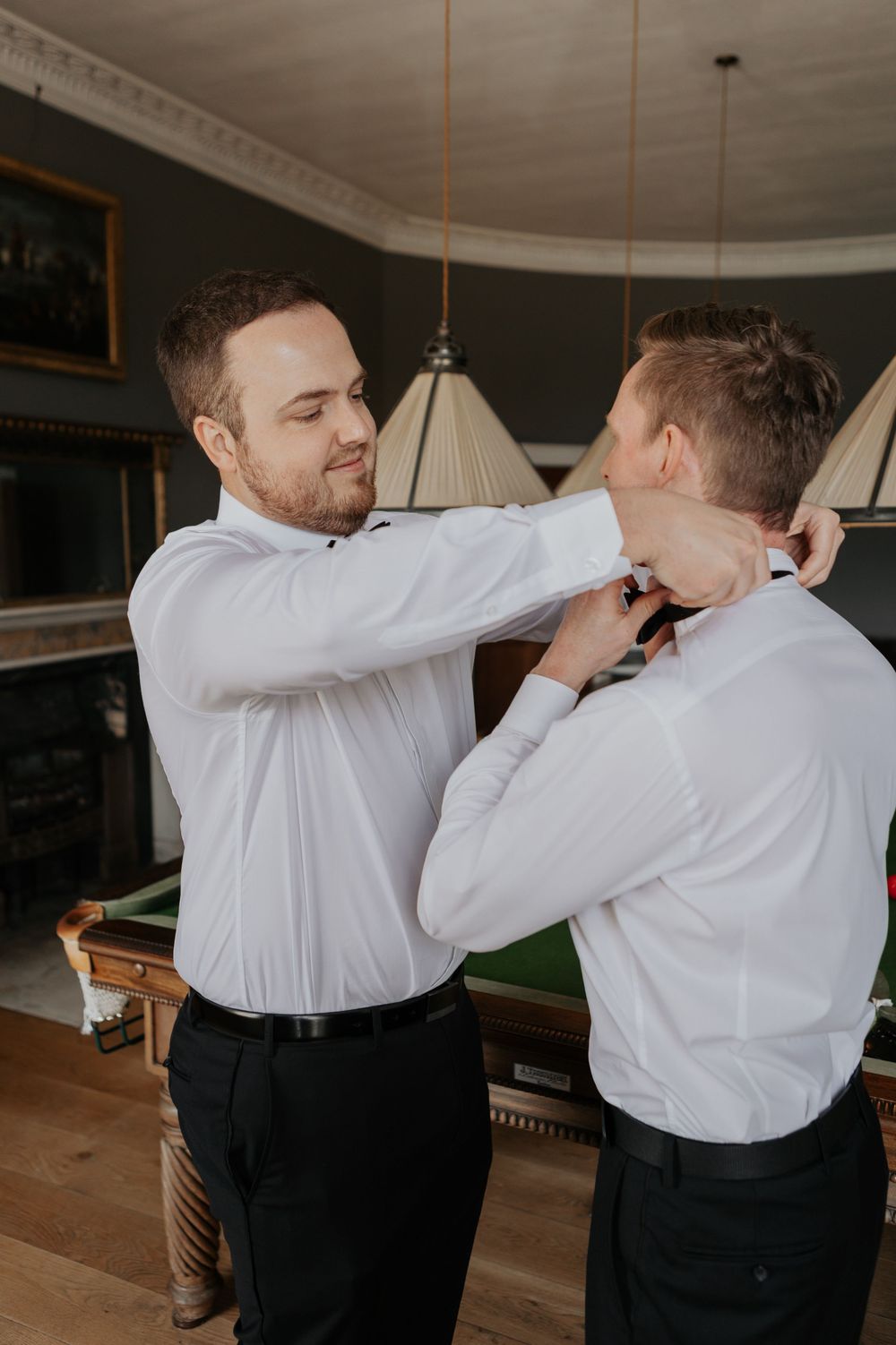 Groom in white shirt smiling while groomsman adjusts his bow tie near a snooker table.