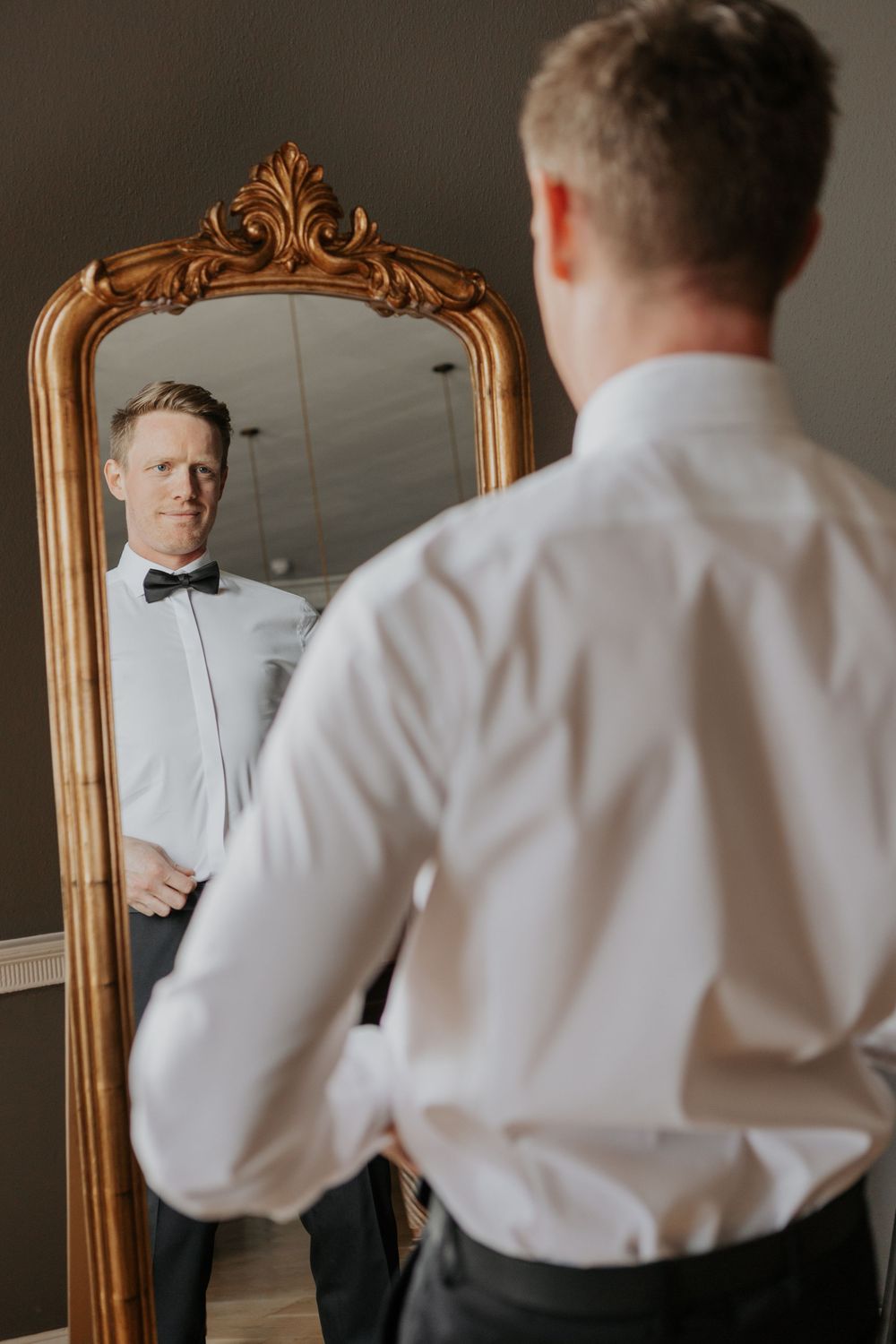 Groom in black tuxedo and bow tie checking his appearance in an ornate gold-framed mirror.