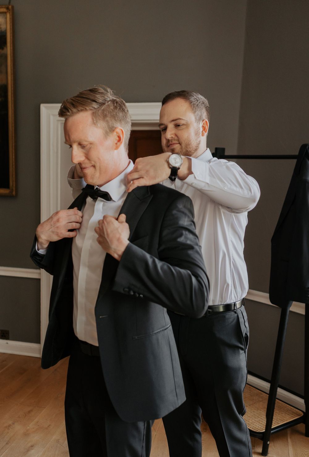 Groomsman in white shirt helping groom put on dark suit jacket before wedding ceremony.