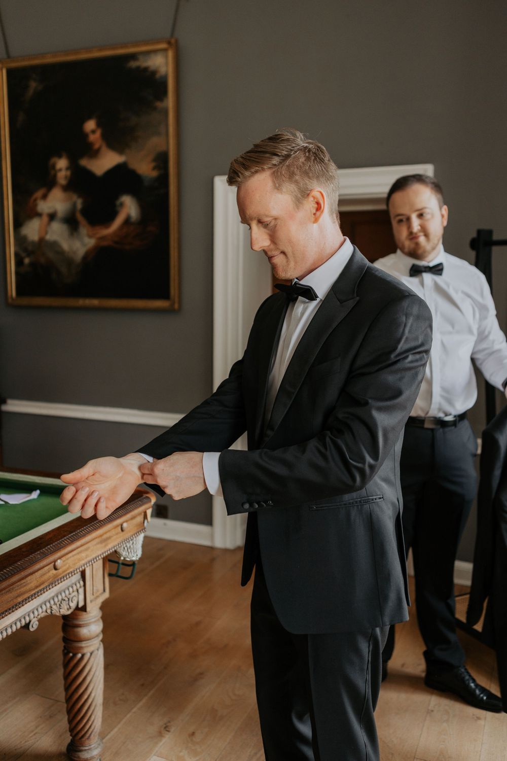 Groom in black tuxedo adjusting watch at billiard table with smiling groomsman in background.