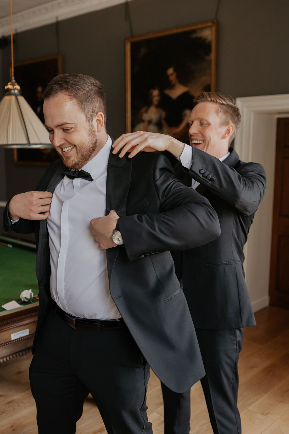 Two men in dark suits laughing while one helps the other into his jacket in elegant room.