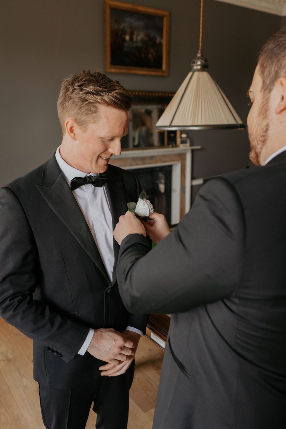 Groomsman pinning white rose boutonniere onto smiling groom's dark suit lapel before wedding.