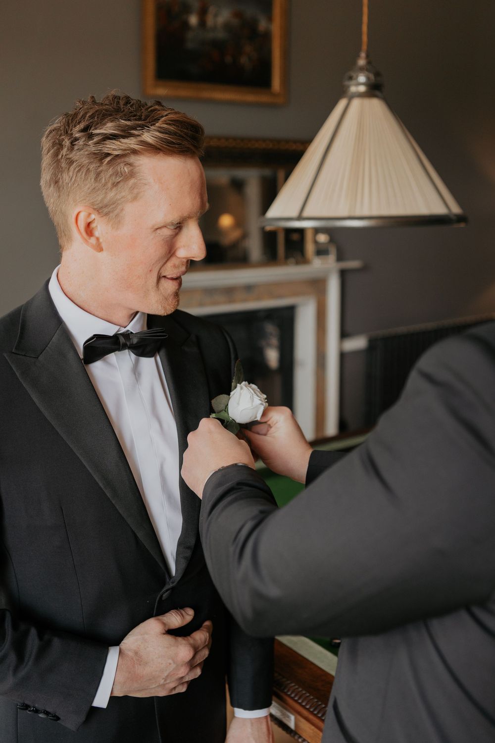 Groomsman carefully attaching white rose boutonniere to groom's dark tuxedo lapel at wedding.