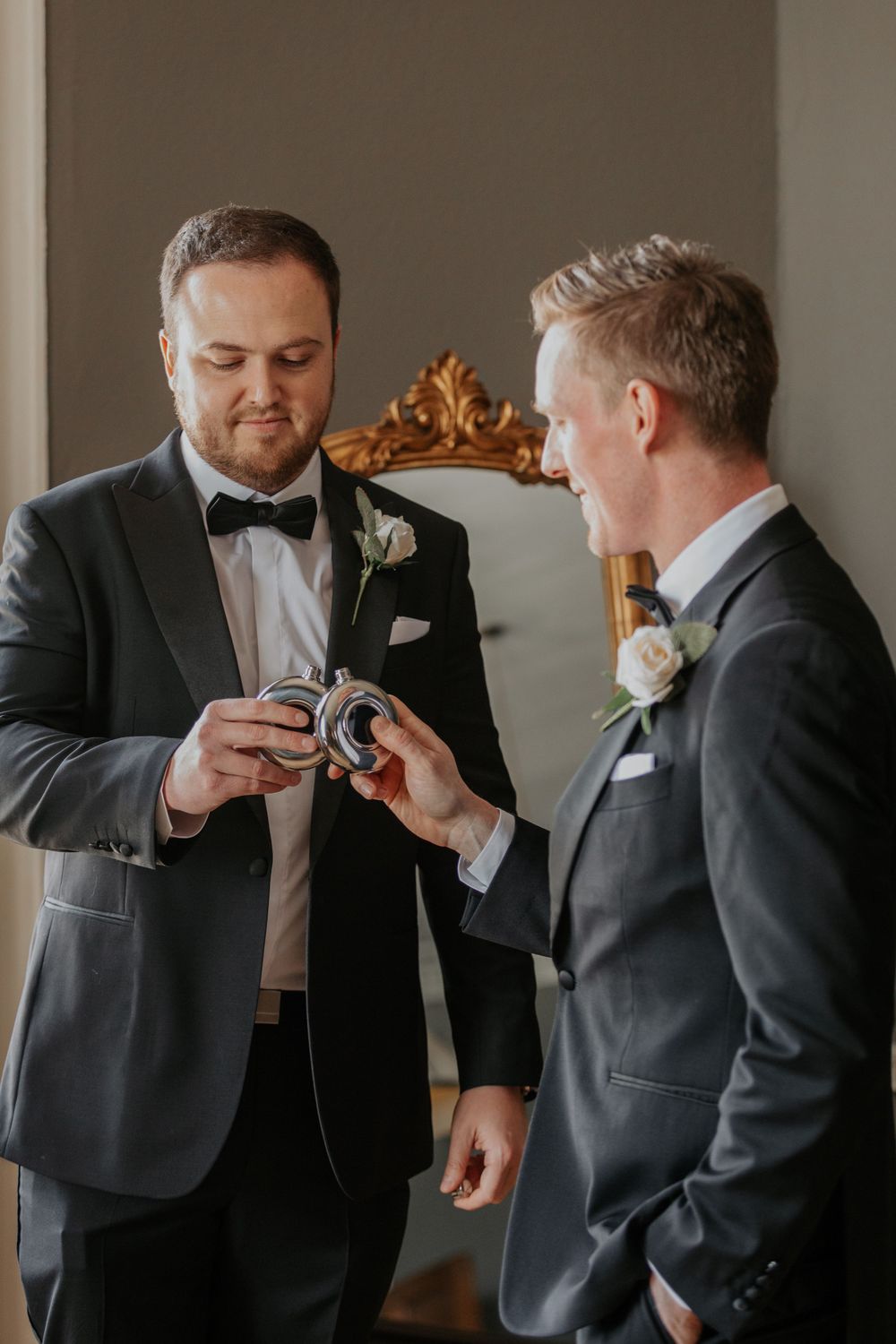 Two grooms in grey suits exchange wedding rings during pre-ceremony preparations in an elegant room.