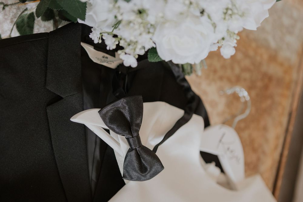 Black tuxedo jacket with a dark grey bow tie displayed beside a white floral wedding bouquet.