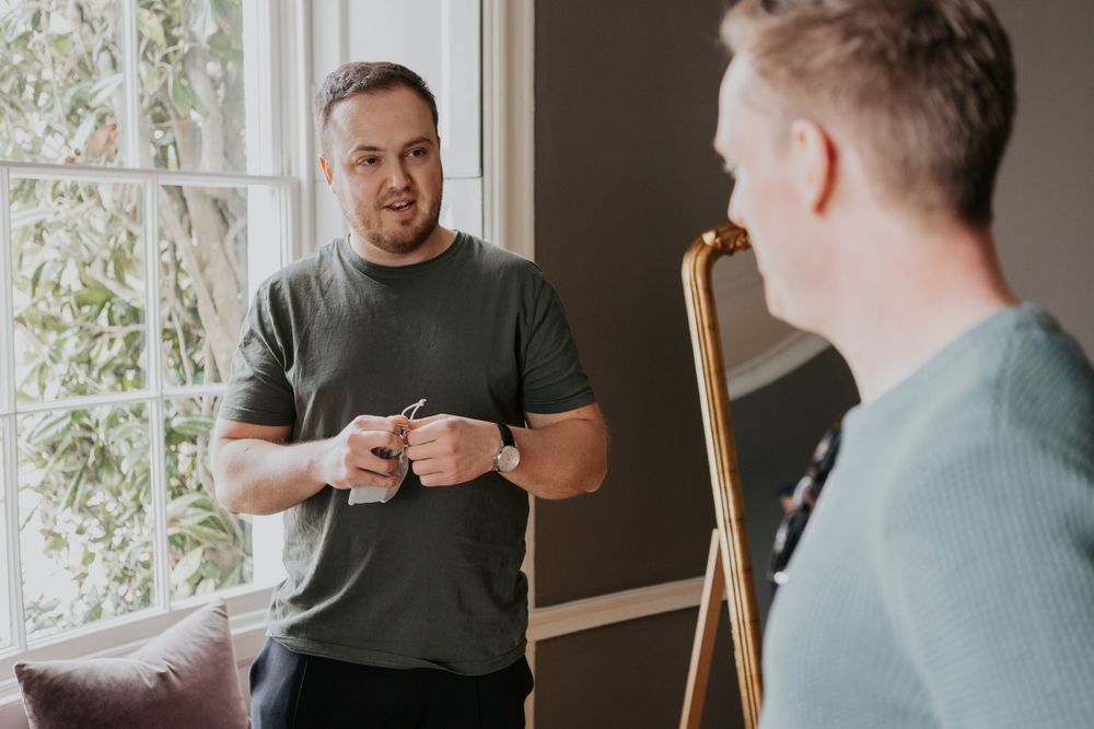 Two men in casual attire having a conversation near a large ornate gold-framed mirror indoors.