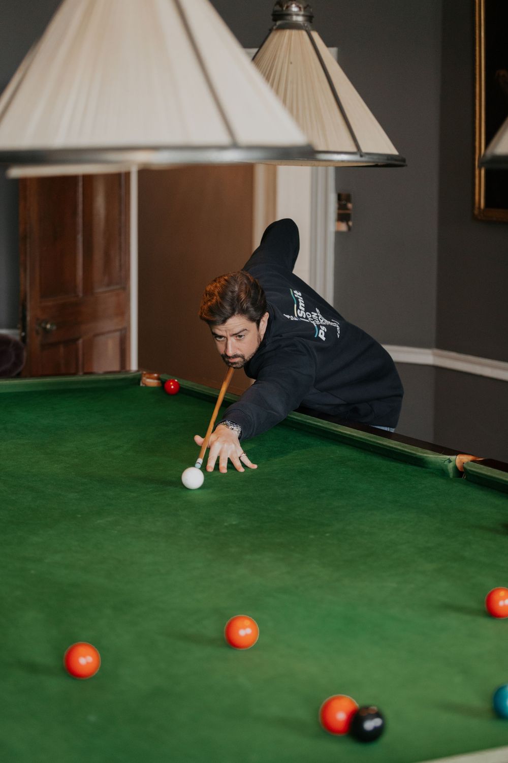 Man in black hoodie leaning over green snooker table taking a shot with cue ball during game.