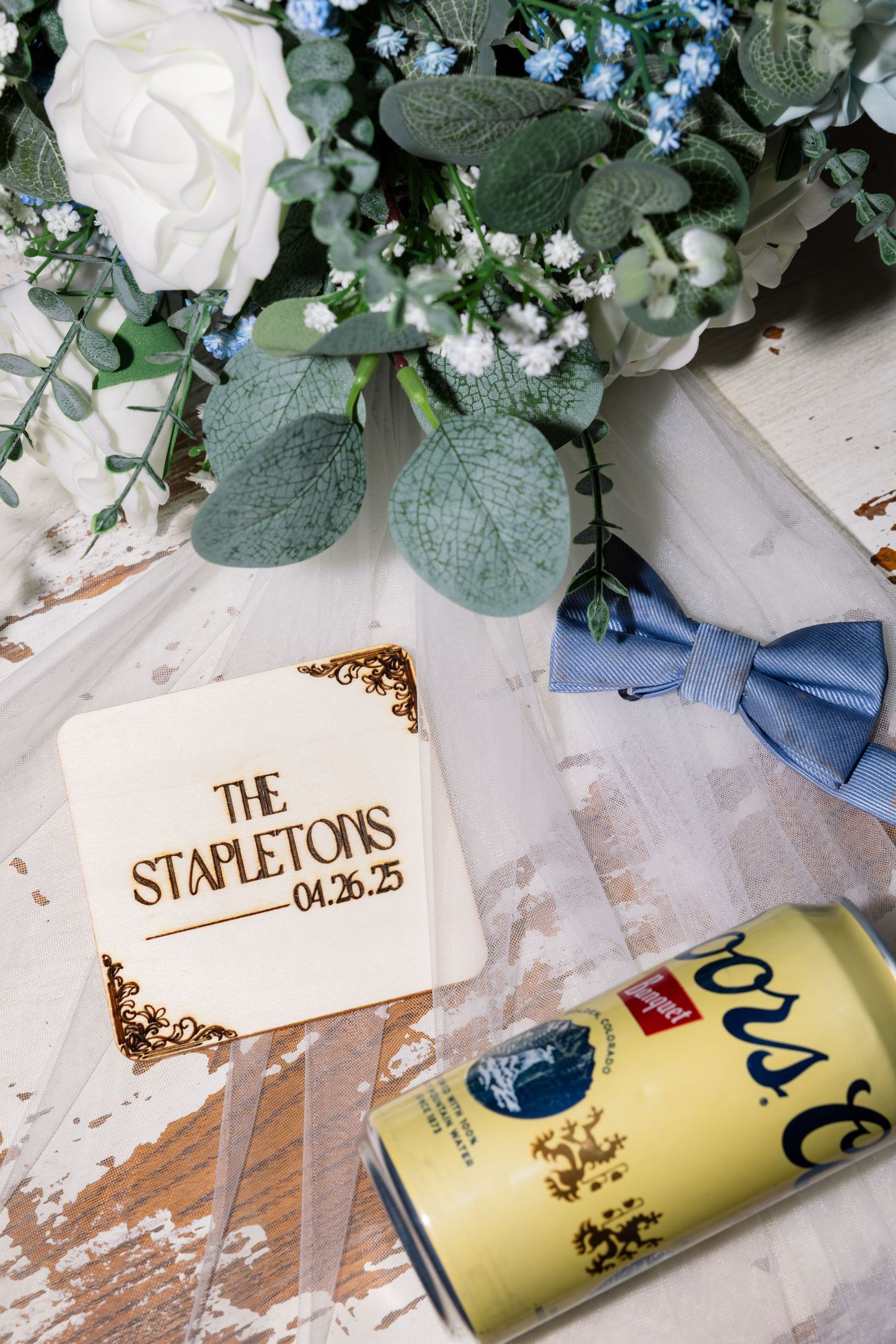 Elegant wedding decor featuring eucalyptus leaves, white flowers, blue bow ties, and yellow beverage cans on a rustic surface.