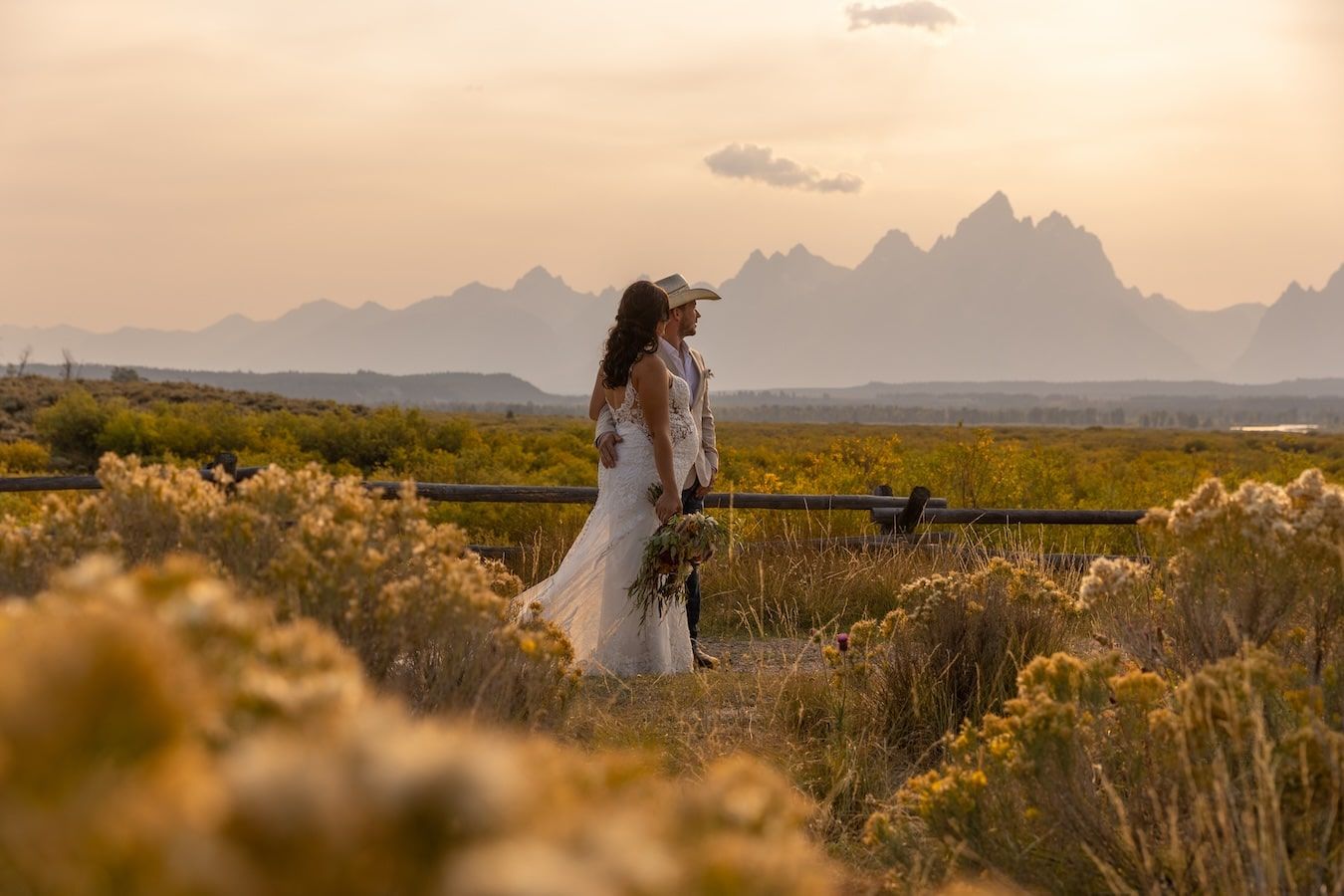A couple stands together in a golden desert landscape at sunset with mountains in the background.