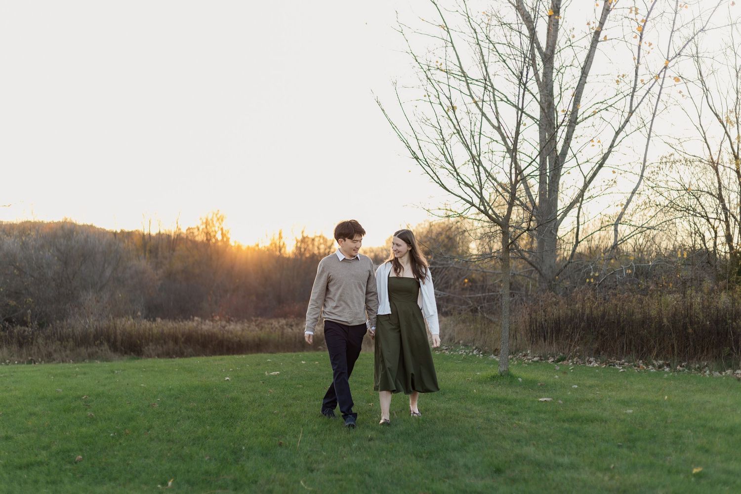 Two people walk hand in hand through a grassy field at sunset while their dog follows behind them.