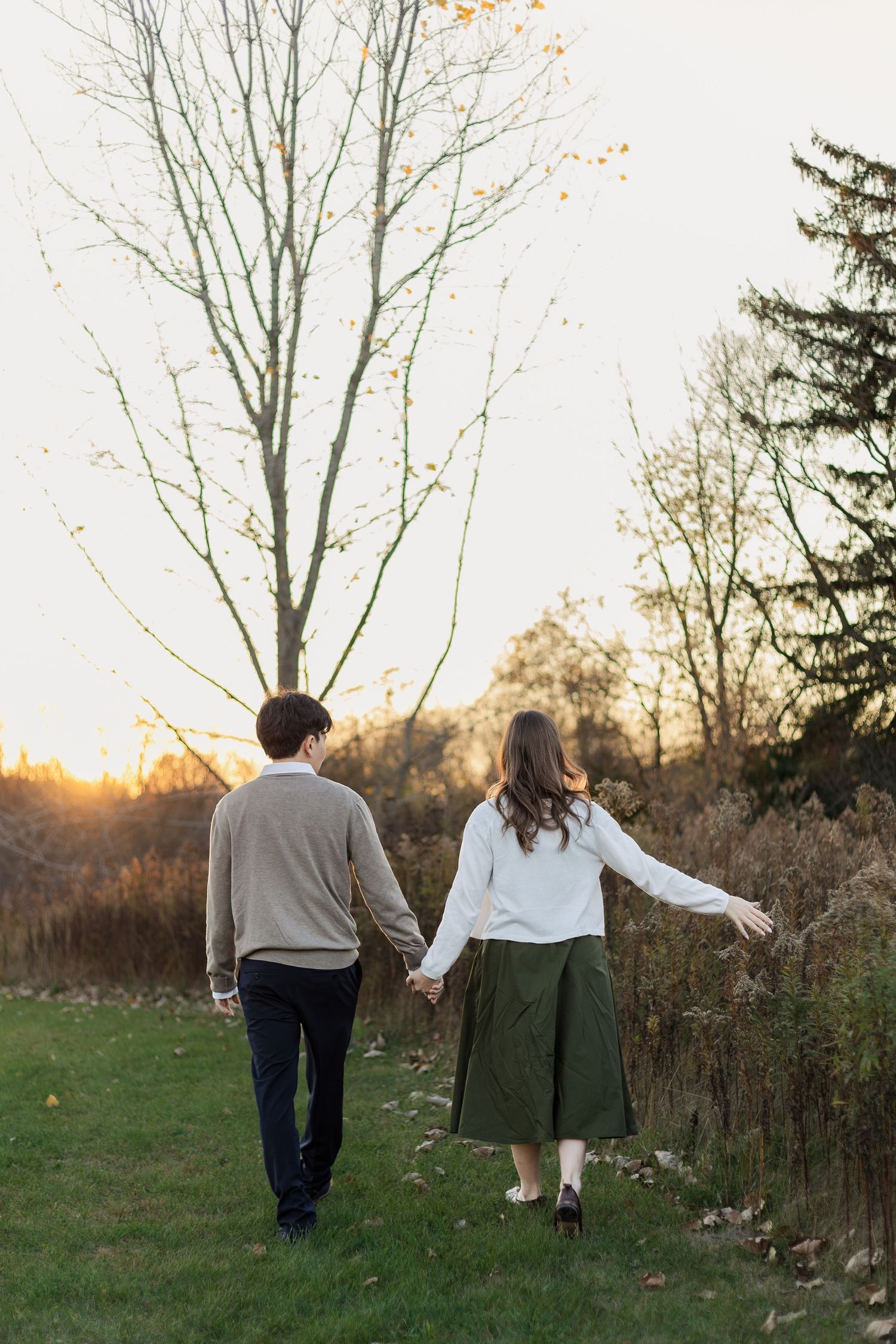 A couple holds hands while walking through a field at sunset with bare trees in the background.