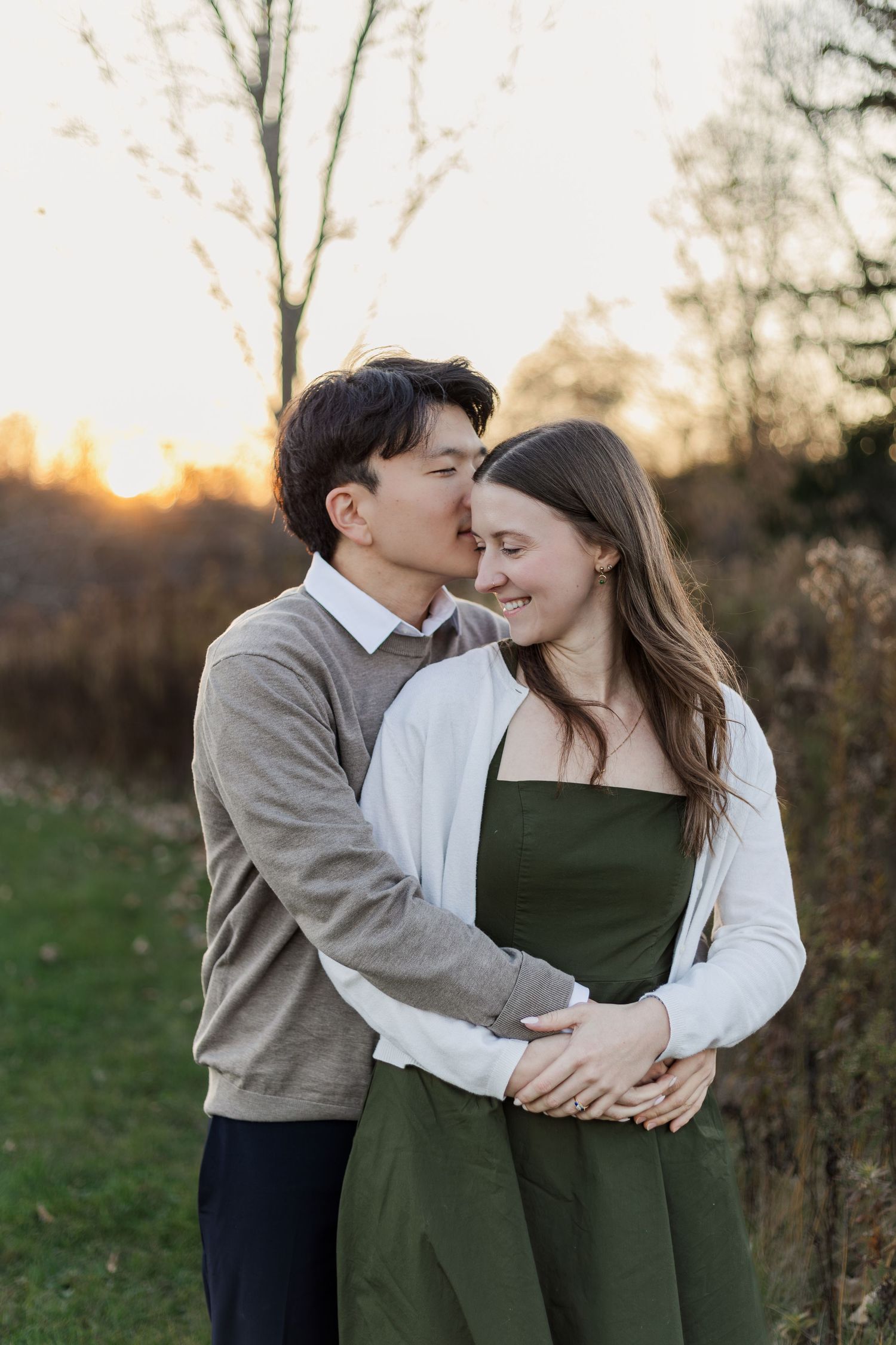 A romantic couple embraces at sunset in a natural outdoor setting with golden light filtering through trees.
