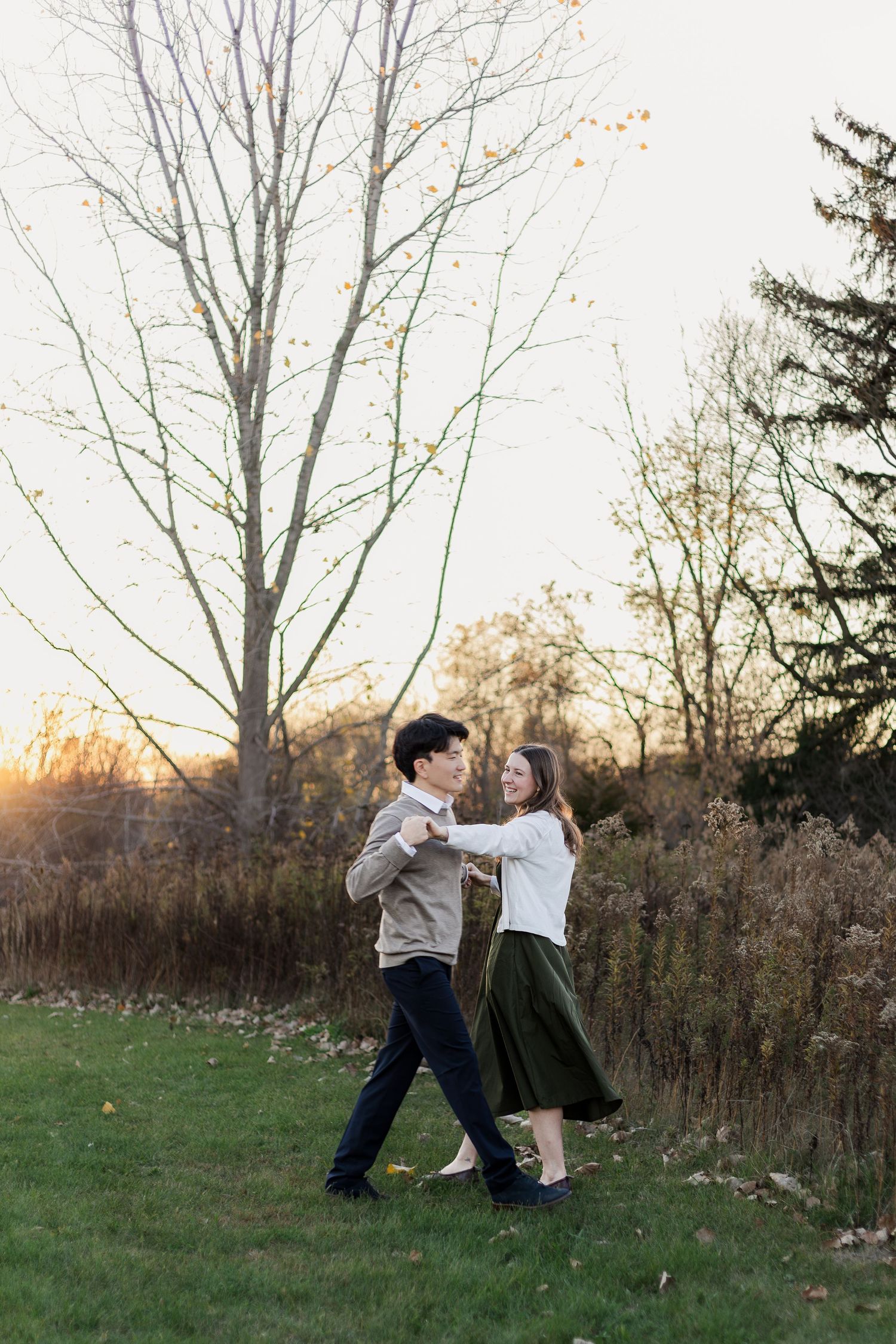 A couple dances together on a grassy lawn at sunset with bare trees and evergreens in the background.