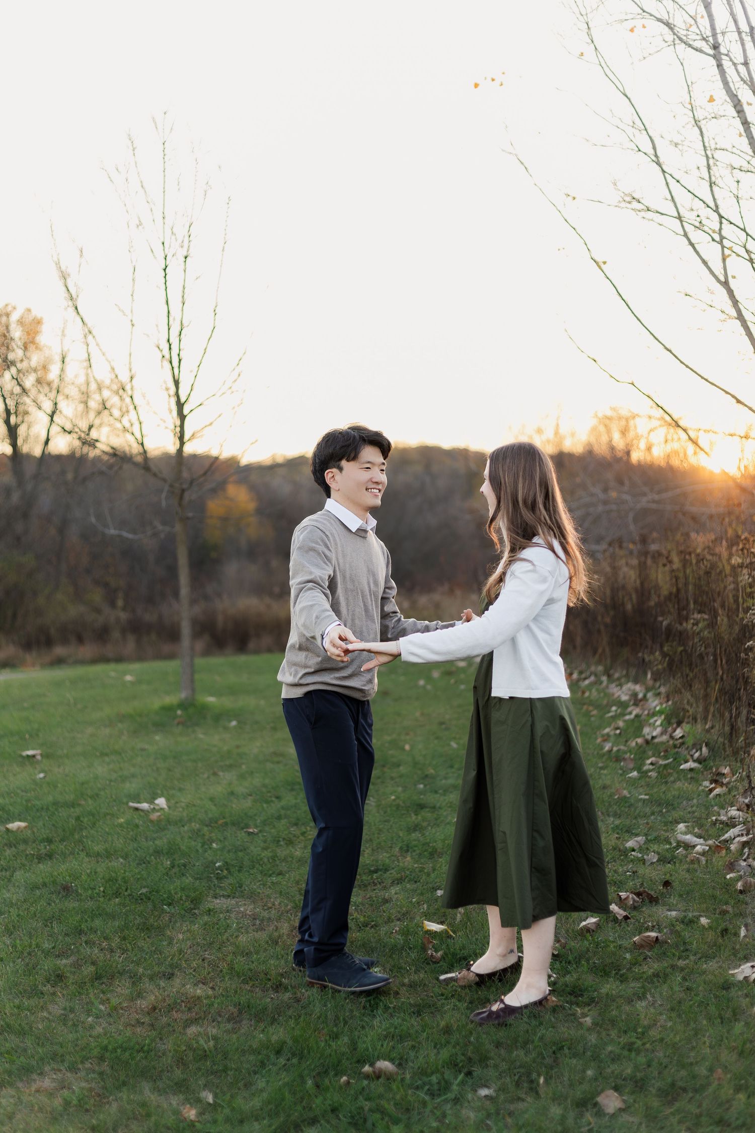 A couple holds hands in a romantic outdoor setting at sunset with autumn trees and grass in the background.