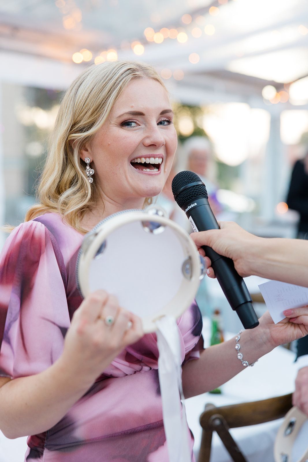 A joyful person in a pink dress laughs while holding a plate and speaking into a microphone at an event.