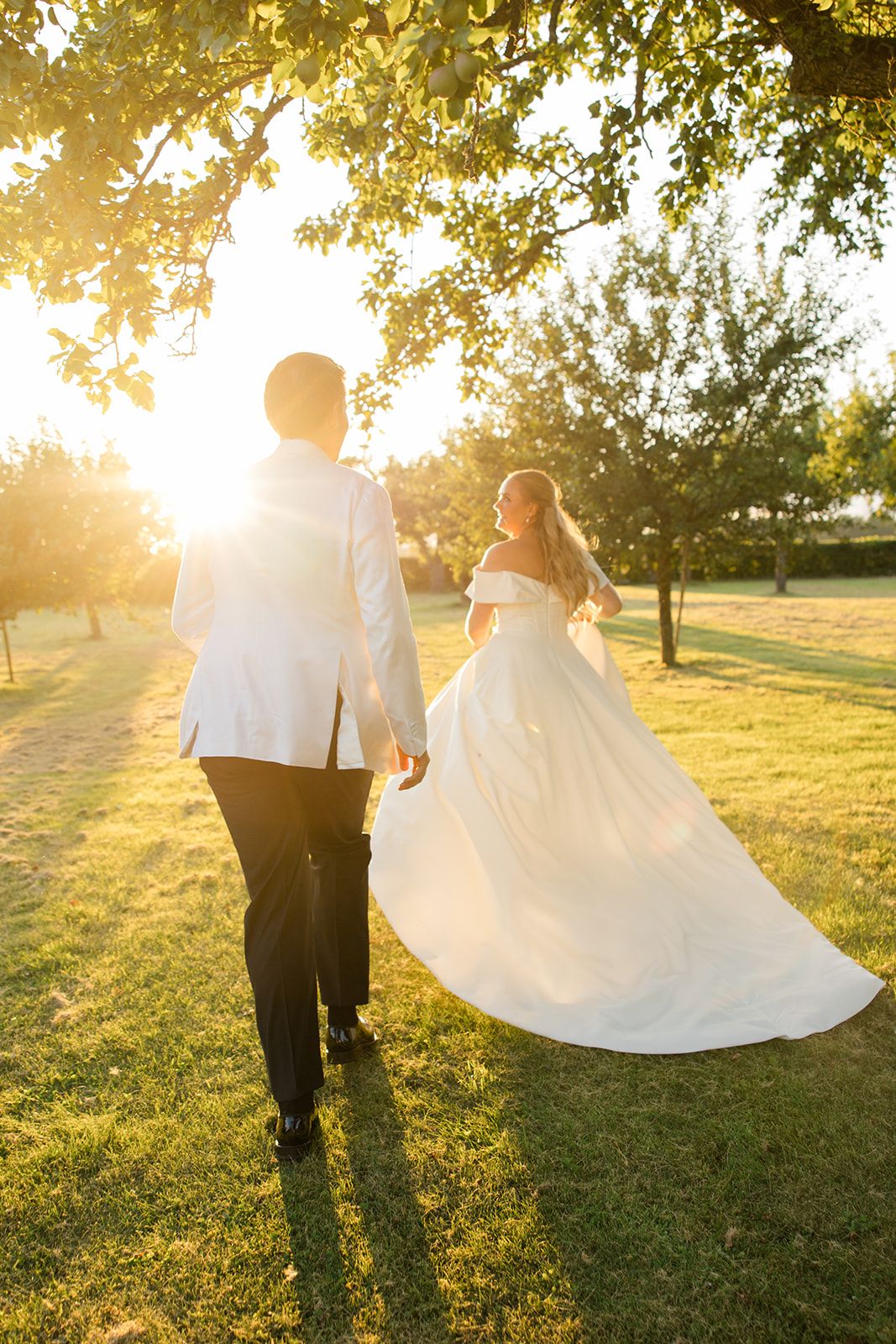 A couple in wedding attire walks through a sunlit meadow during golden hour as the bride's white dress flows behind them.