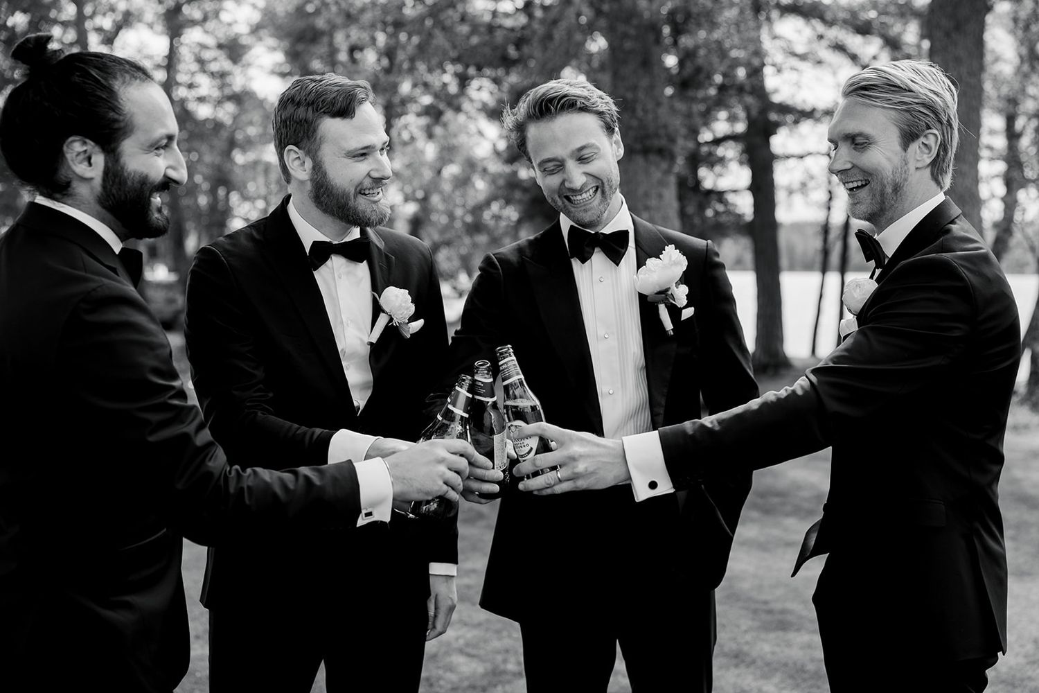 Groomsmen in black tuxedos share drinks and laughs during an outdoor wedding celebration.