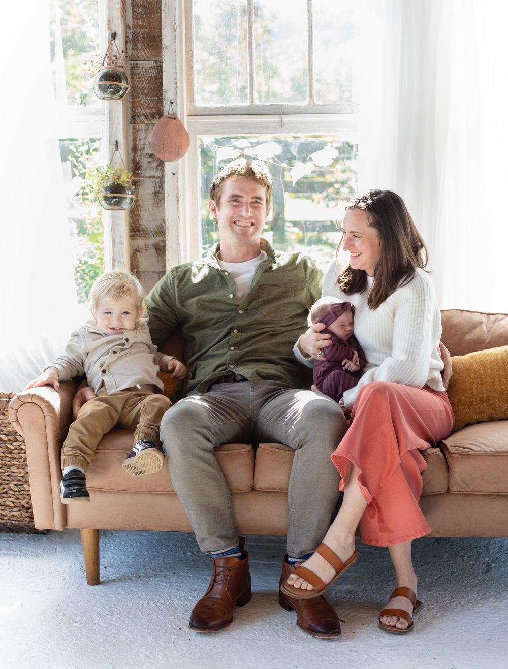 A family relaxes together on a tan leather couch in a bright sunlit room with large windows.