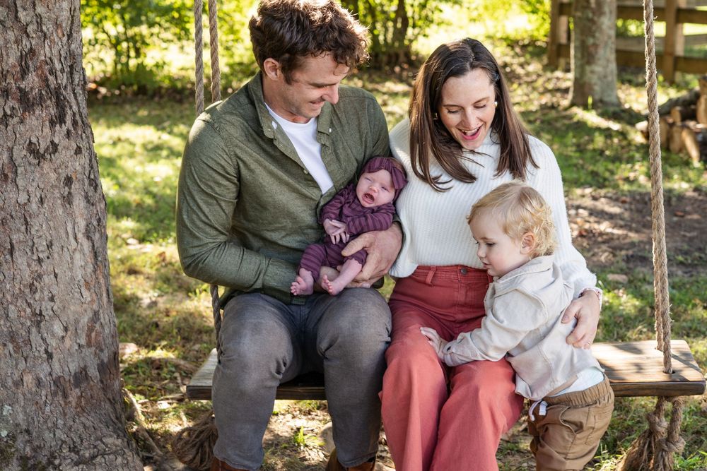 A family sits together outdoors on a sunny day surrounded by trees in a natural wooded setting.