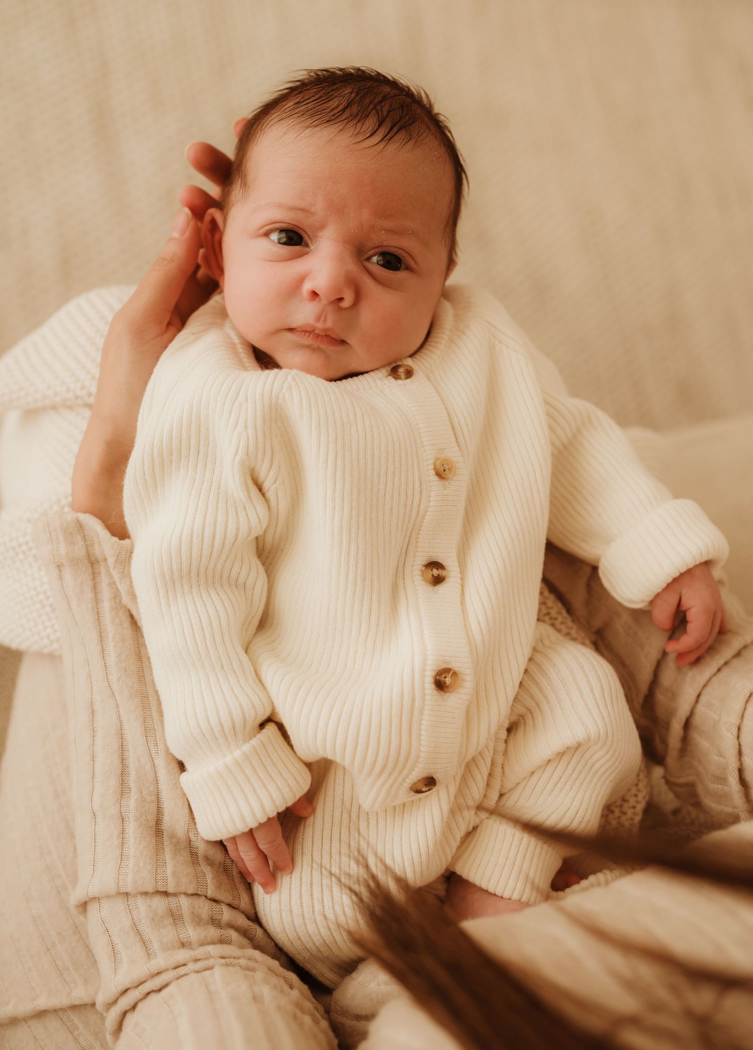 An infant wearing a cozy cream-colored ribbed cardigan lounges on soft beige cushions in warm lighting.