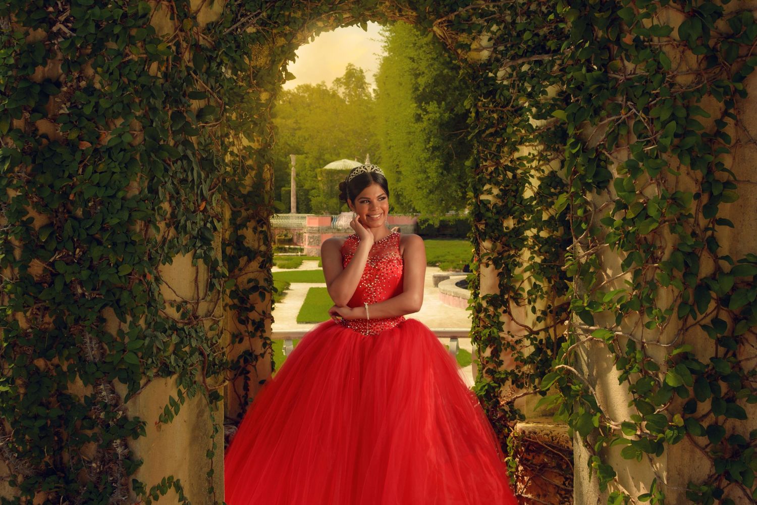 Red ballgown framed by garden archway during golden hour.