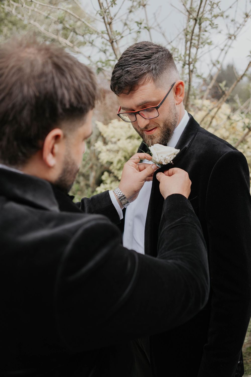 Groomsman pinning a white boutonniere onto a bearded groom in a black velvet suit outdoors.