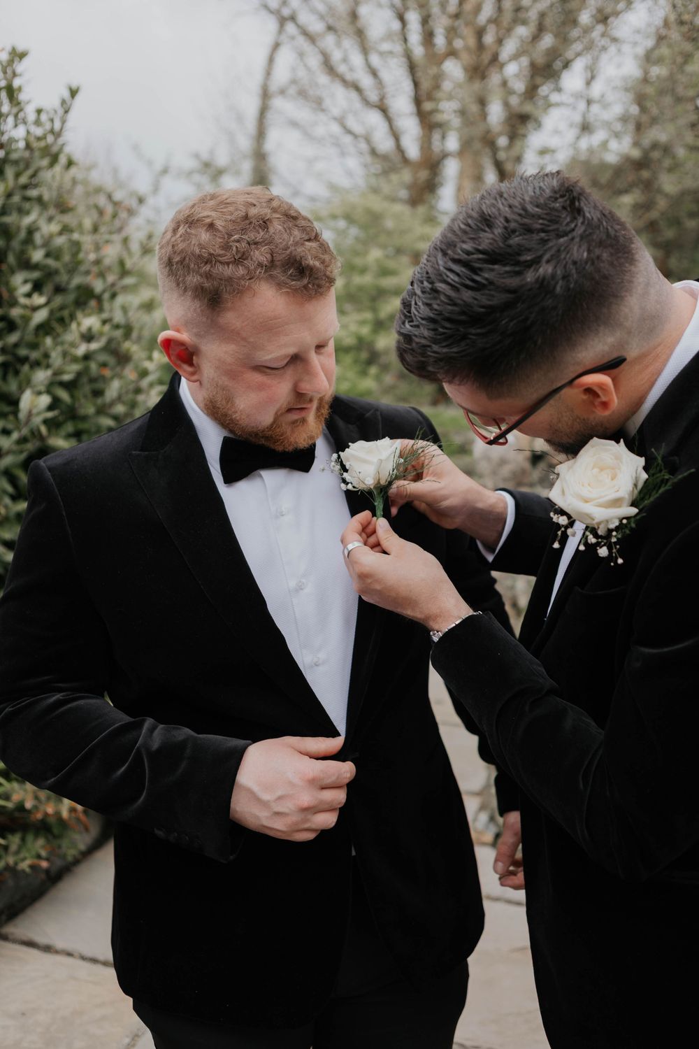 Man in black tuxedo with bow tie having a white floral boutonniere pinned to his lapel outdoors.