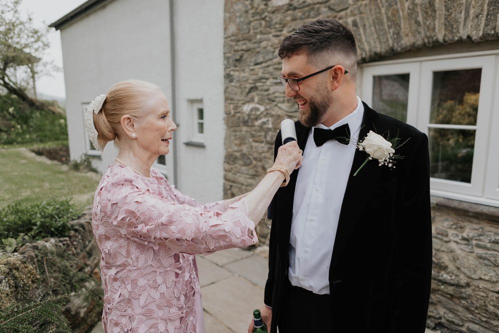 Woman in pink floral dress adjusting boutonniere on groom in black tuxedo outside stone building.