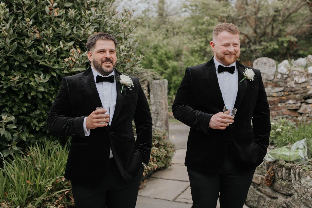 Two groomsmen in black velvet tuxedos with bow ties walking together holding drinks outdoors.