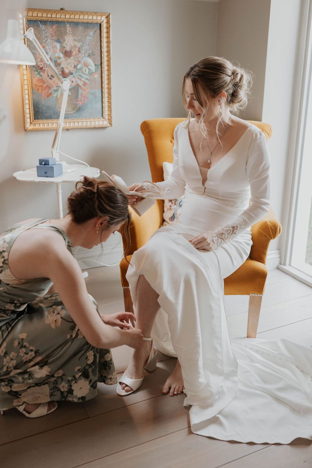Bridesmaid in floral dress helping bride put on wedding shoes while seated in yellow chair indoors.