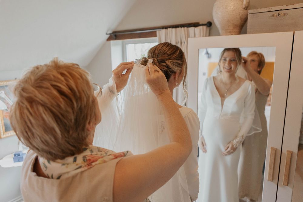 Mother helping bride with veil in bright dressing room, bride reflected in mirror wearing white gown.