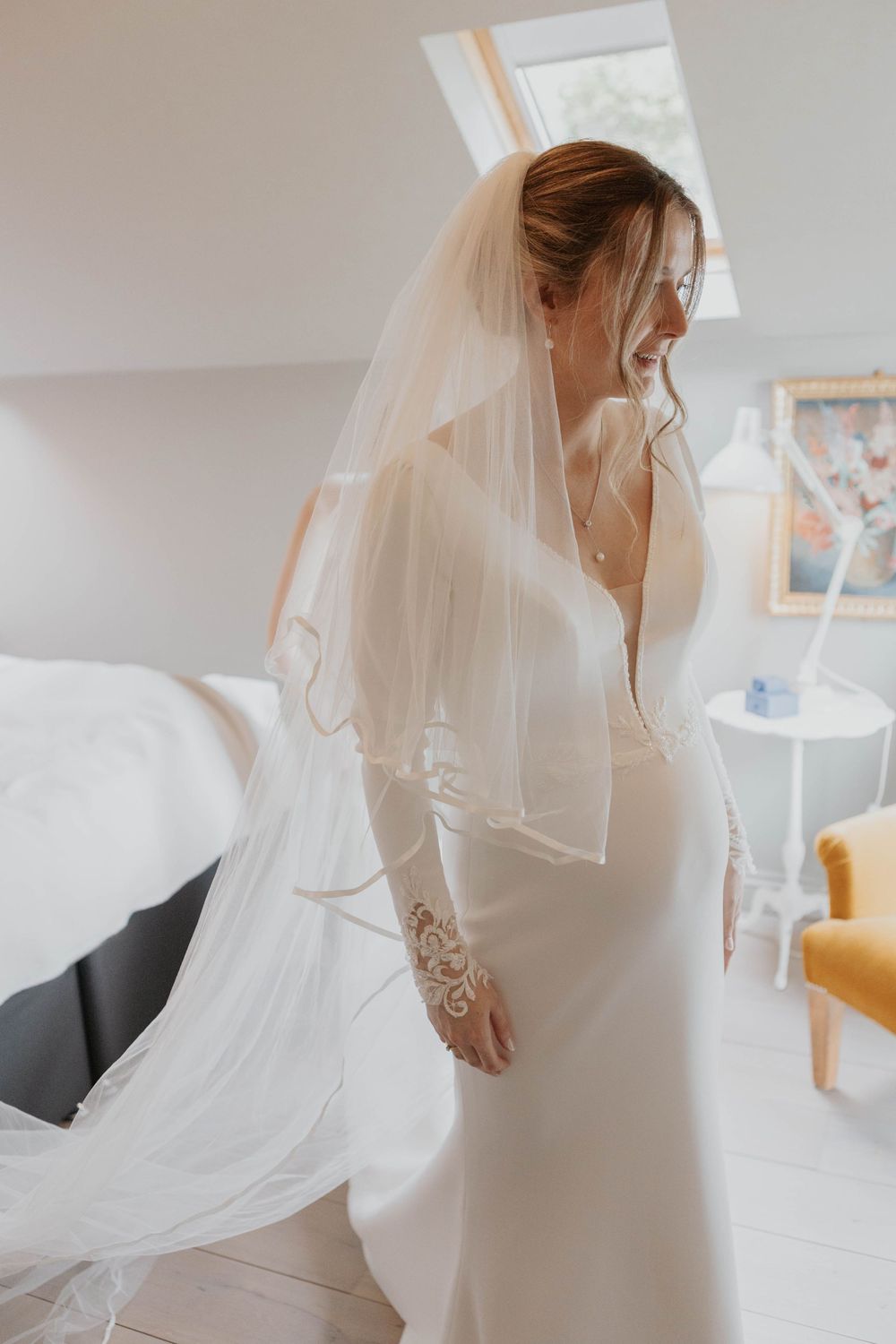 Bride in elegant white long-sleeve wedding dress and layered veil posing in bright dressing room.