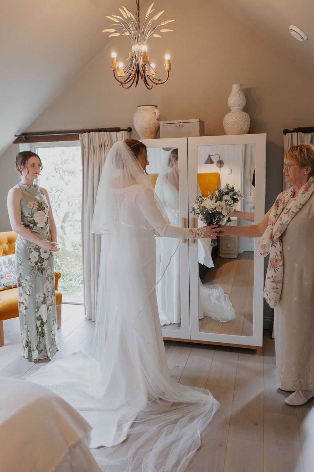 Bride in white gown and veil holding bouquet before mirror with bridesmaid and mother nearby.