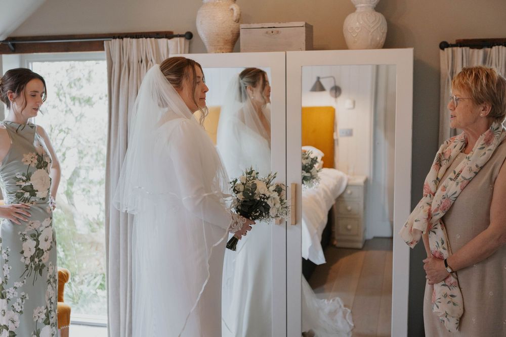 Bride in white gown and veil holding bouquet viewed in mirror with bridesmaid and mother beside her.