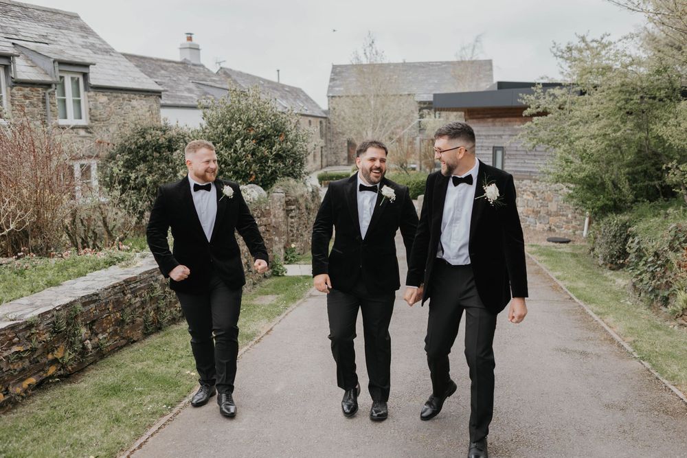 Three groomsmen in black velvet tuxedos with bow ties walking and laughing along an outdoor pathway.