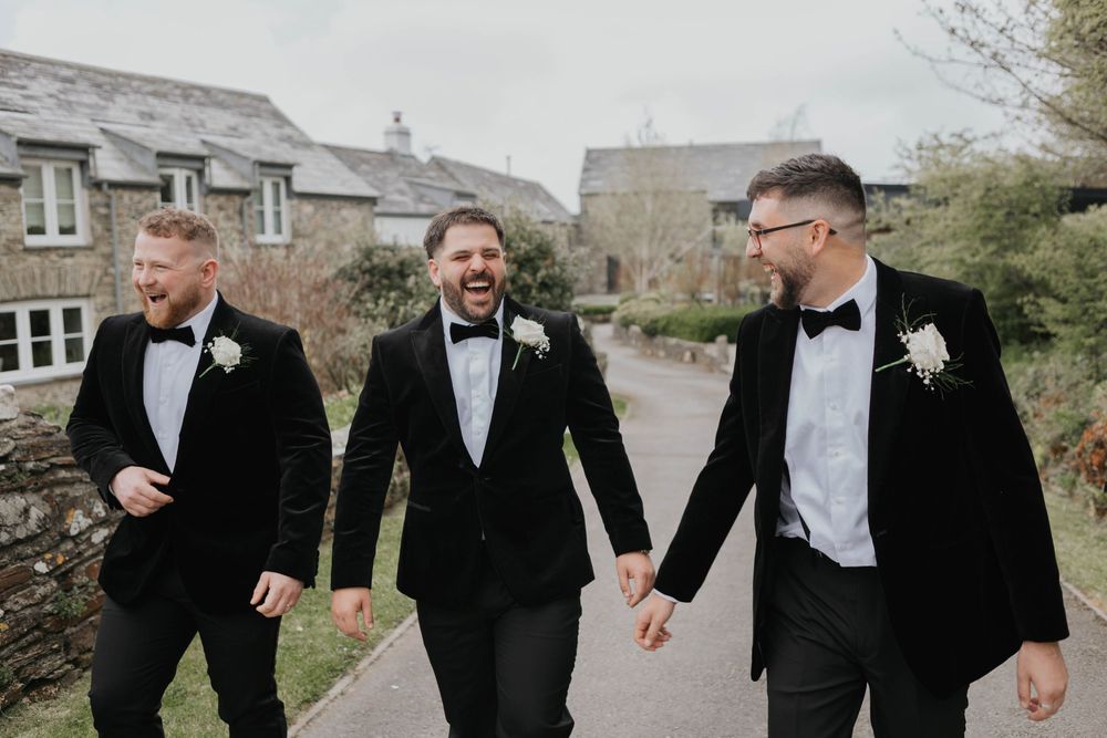 Three groomsmen in black tuxedos with bow ties and white boutonnieres laughing together outdoors at a wedding.