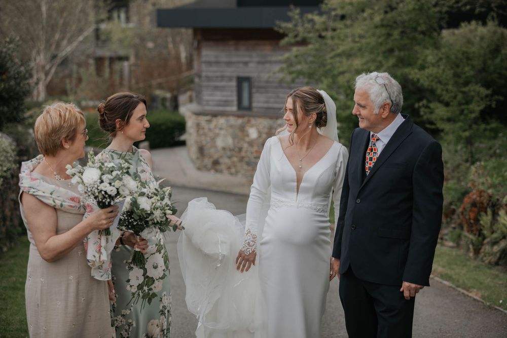 Bride in white wedding dress holding bouquet poses with family members including parents outdoors at wedding venue.