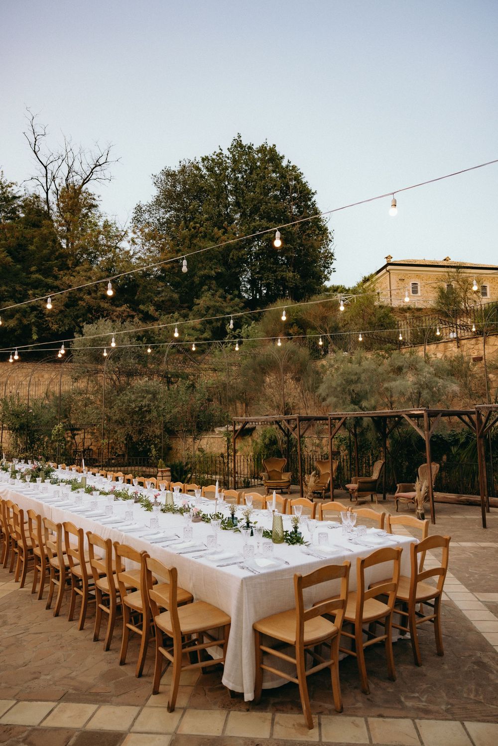 Long banquet tables with white linens and wooden chairs set up outdoors under string lights at dusk.
