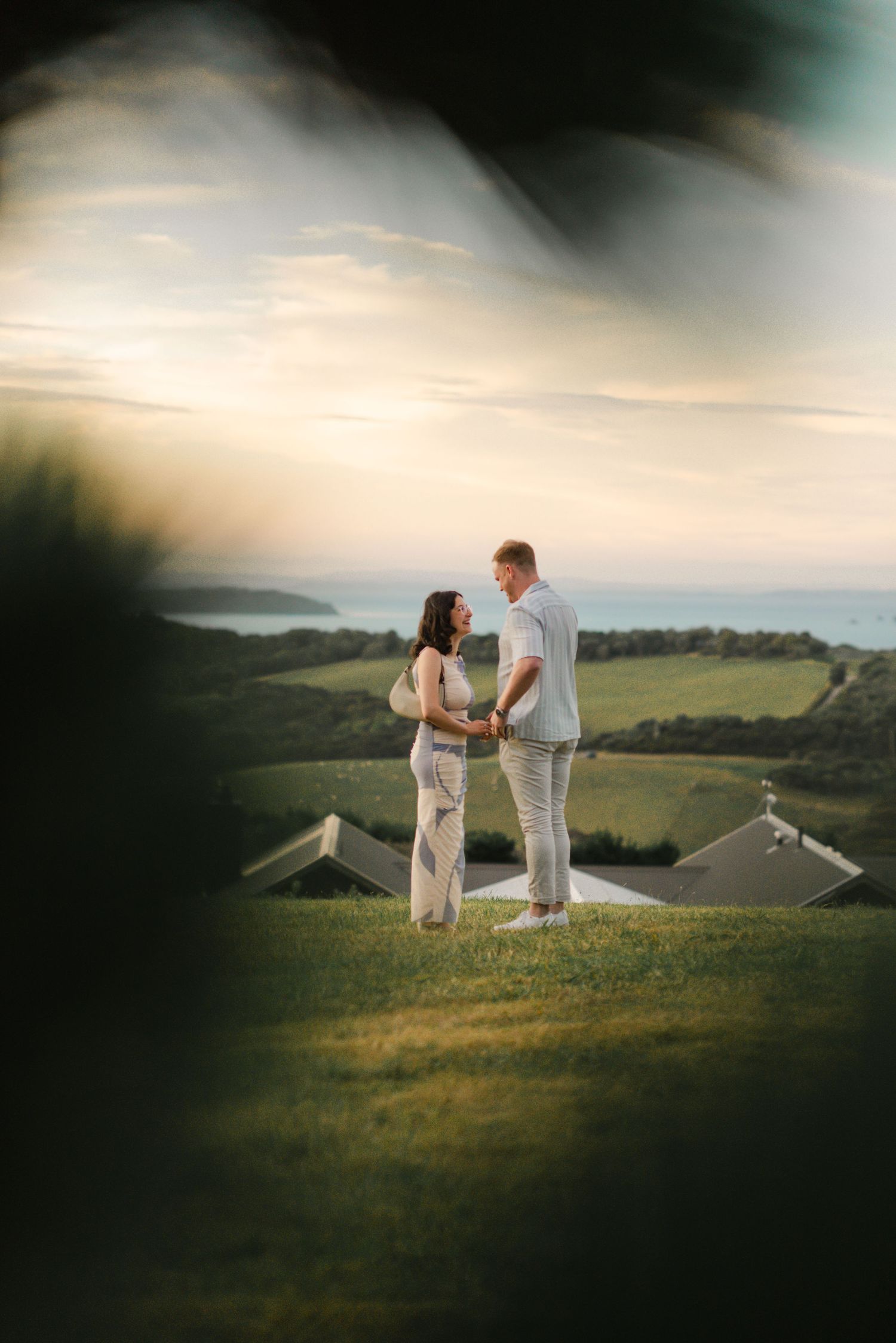 A couple in white and pink attire poses joyfully in a green outdoor setting on a cloudy day.
