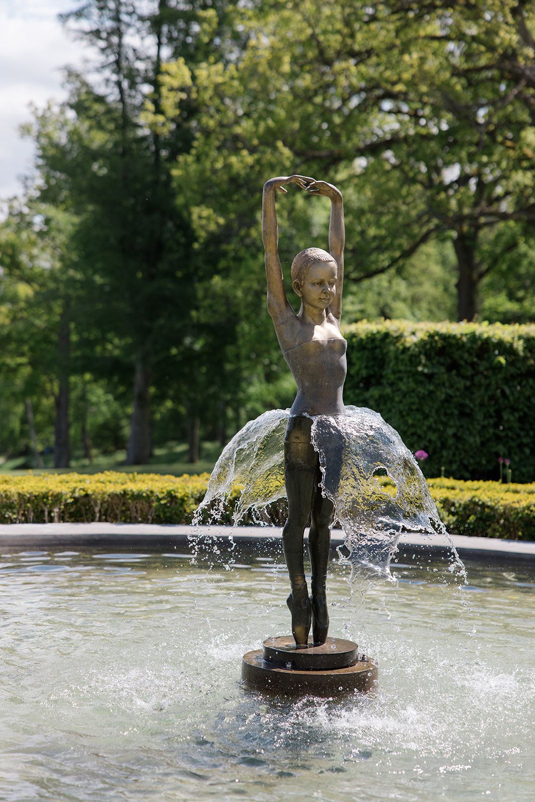 A bronze statue of a child stands in a fountain as water sprays upward creating a dynamic splash effect in a garden setting.