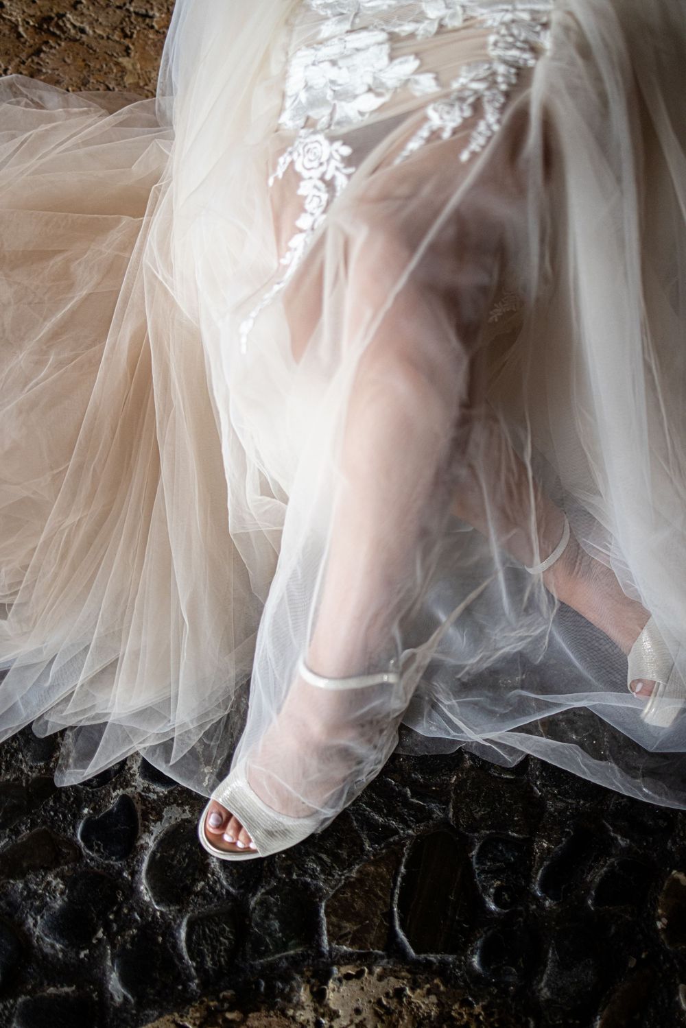 A foot wearing a white peep-toe shoe peeks out from beneath layers of delicate tulle wedding dress fabric.